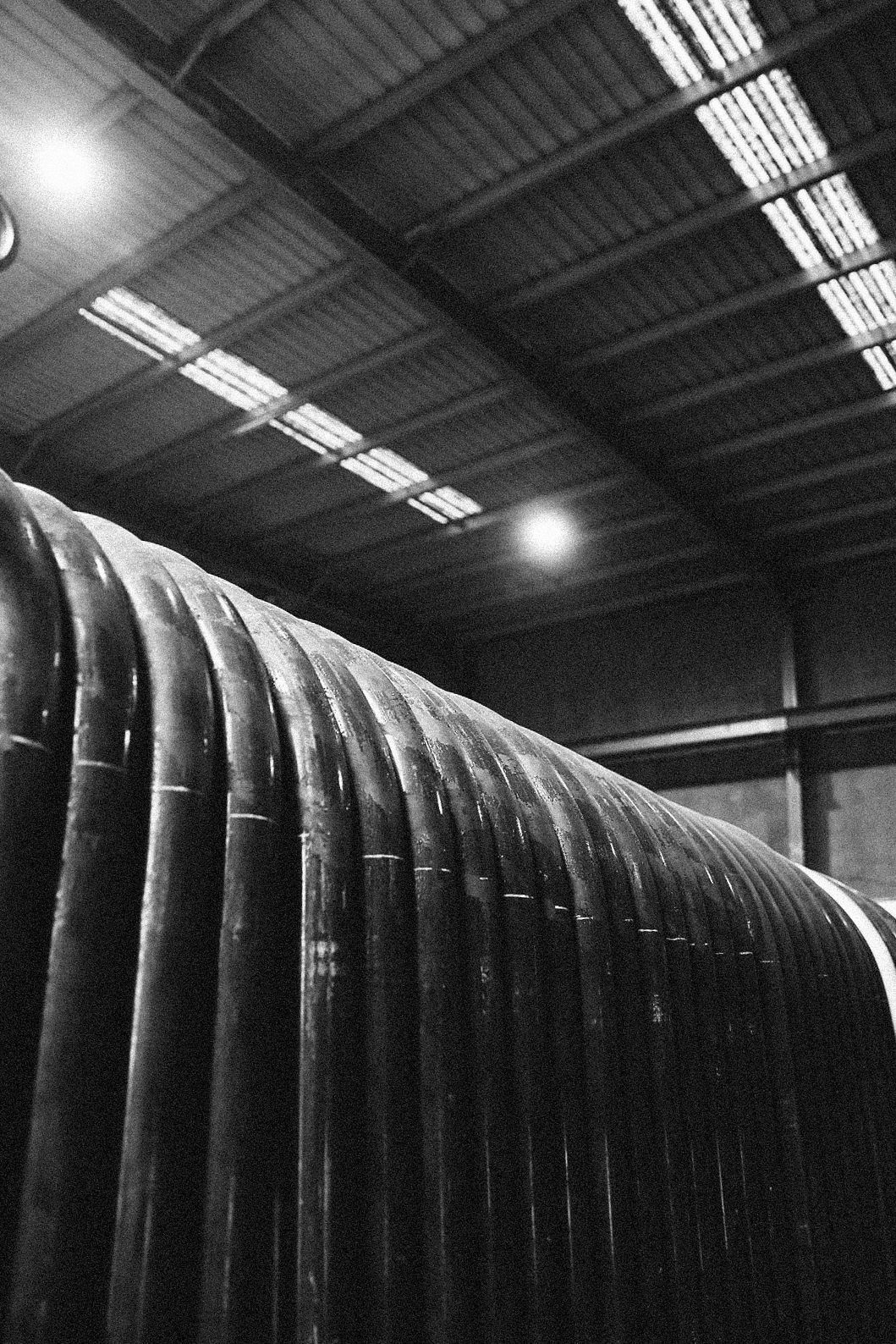 Close-up of numerous dark, cylindrical industrial pipes arranged in a row inside a large, illuminated warehouse.