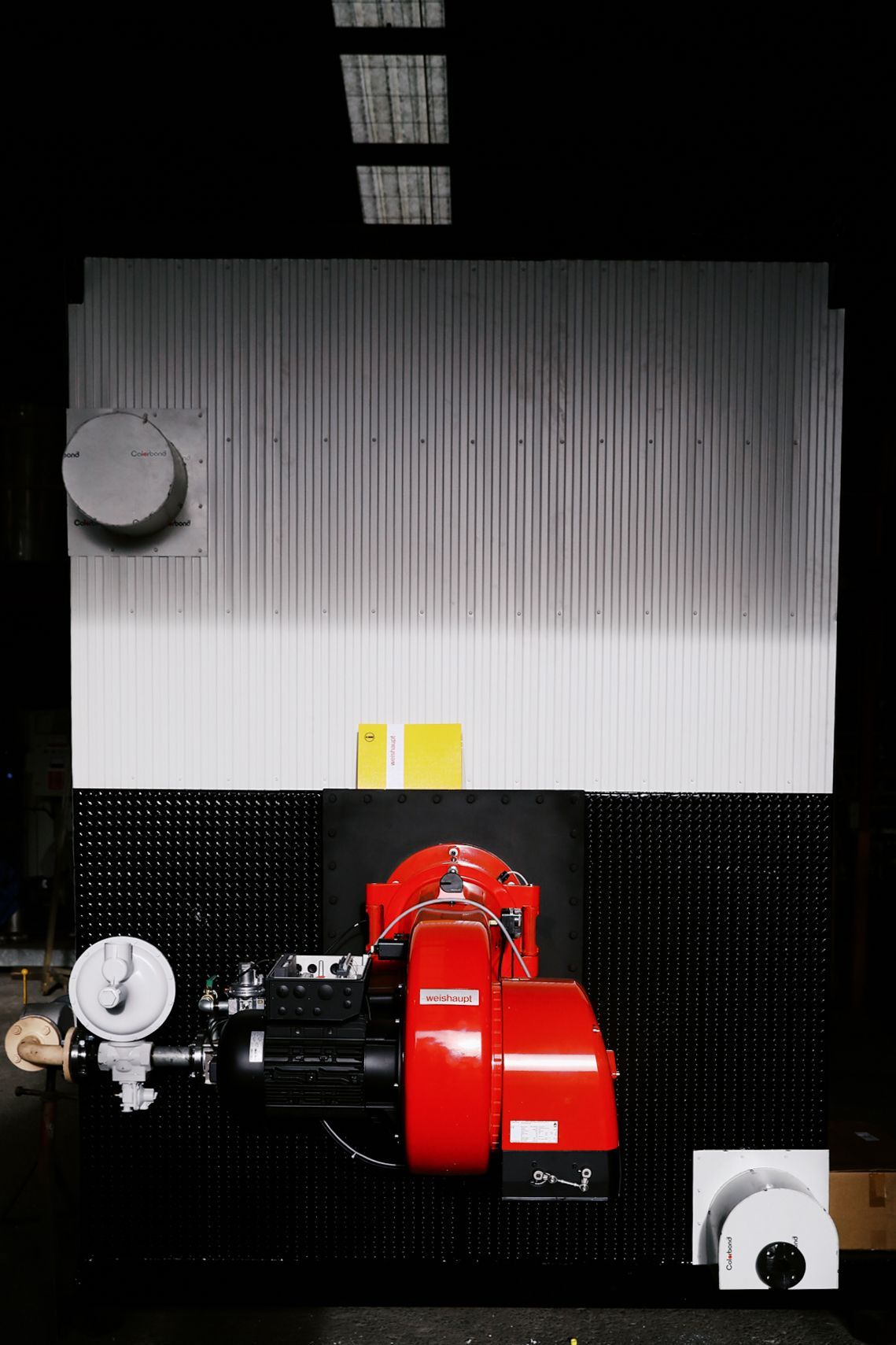 A red commercial burner attached to a large, white industrial boiler unit in a dark room.