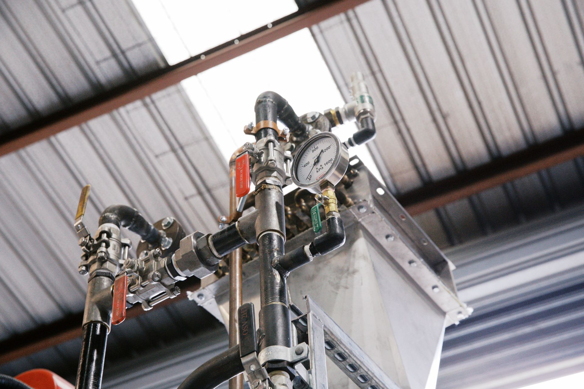 A close-up of industrial machinery featuring black pipes, red-handled valves, and a circular pressure gauge under a roof.
