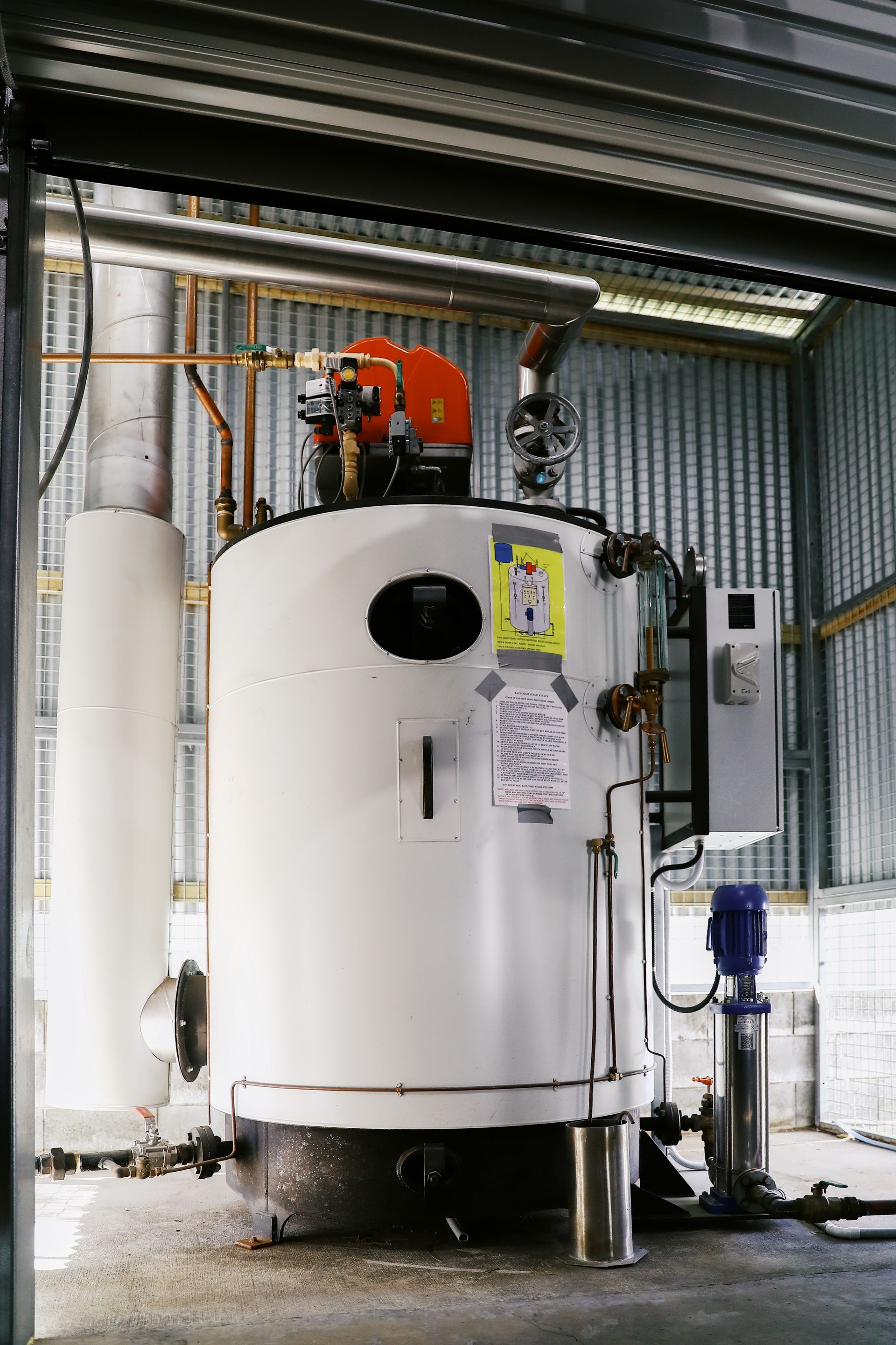 A white industrial boiler sits in a metal shed with a tall exhaust pipe, pump, and control panel attached to its side.