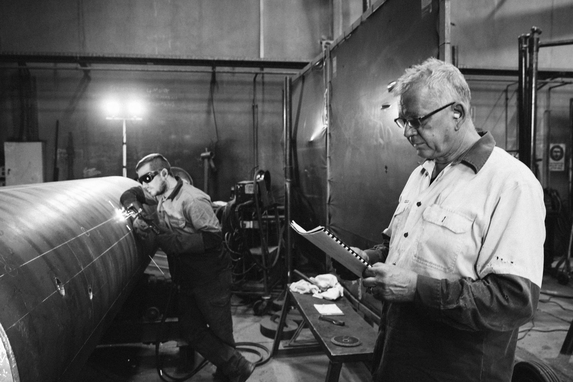 An industrial worker welds a large pipe while a colleague holds a document and monitors the process in a workshop.