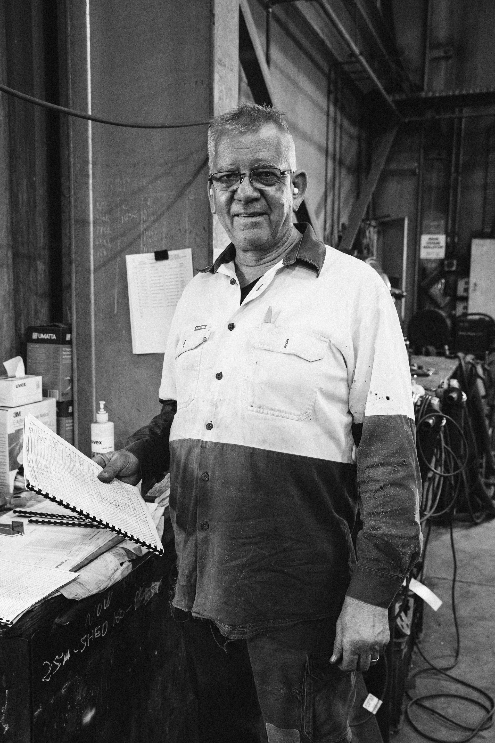 A person in a two-tone work shirt smiles while holding papers in an industrial workshop setting.