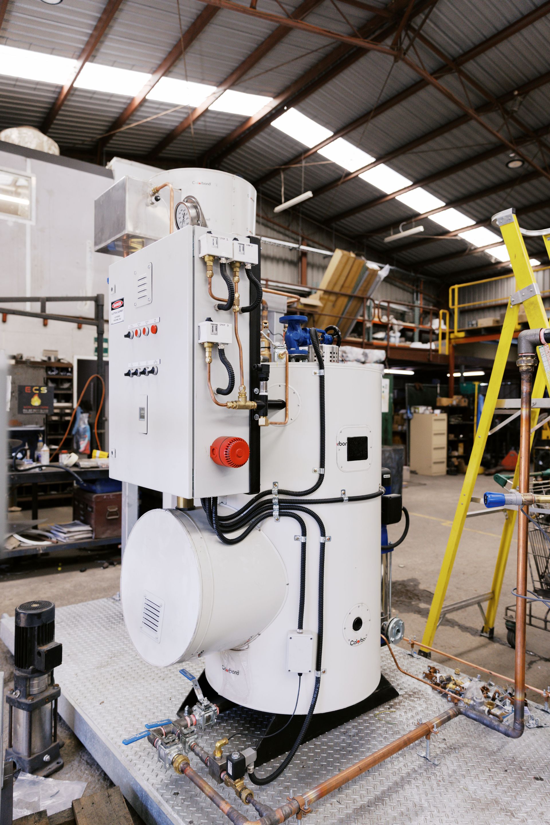 A white industrial steam boiler stands on a platform inside a warehouse with a yellow ladder nearby.