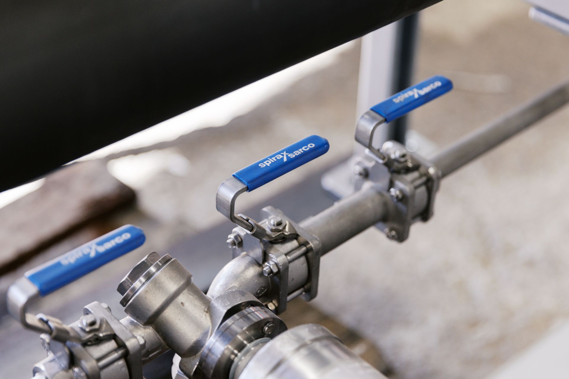 Three stainless steel pipes with blue-handled ball valves mounted on a metal frame.