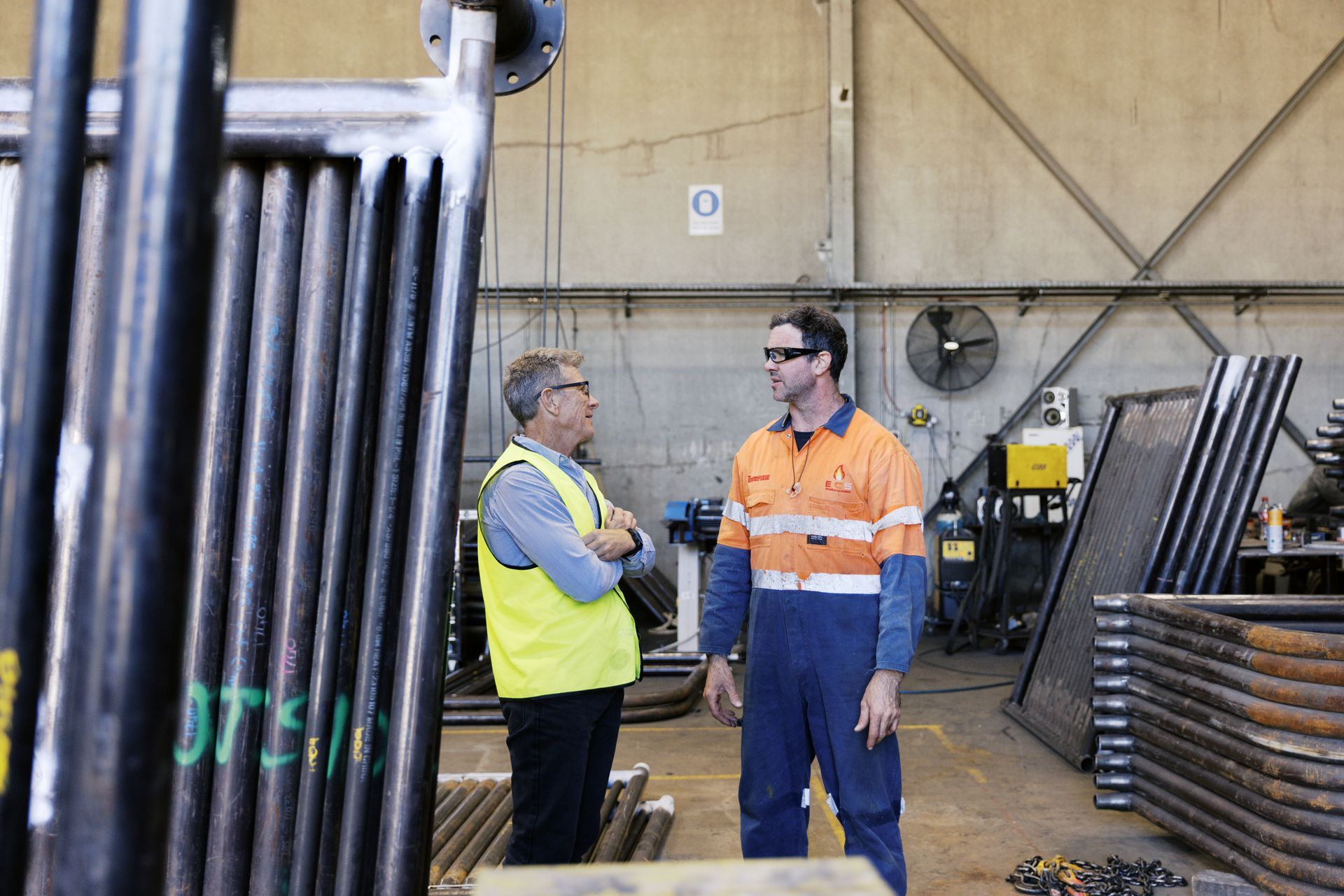Two people in high-visibility work gear talk in an industrial warehouse filled with metal pipe components.