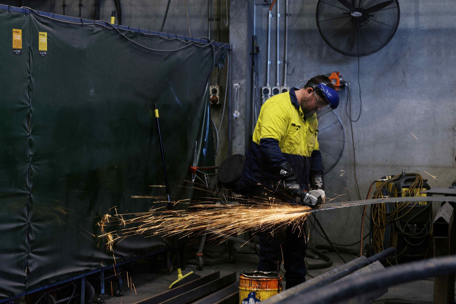 A worker in a yellow and navy high-visibility jacket grinds metal, creating a bright spray of sparks in a workshop.