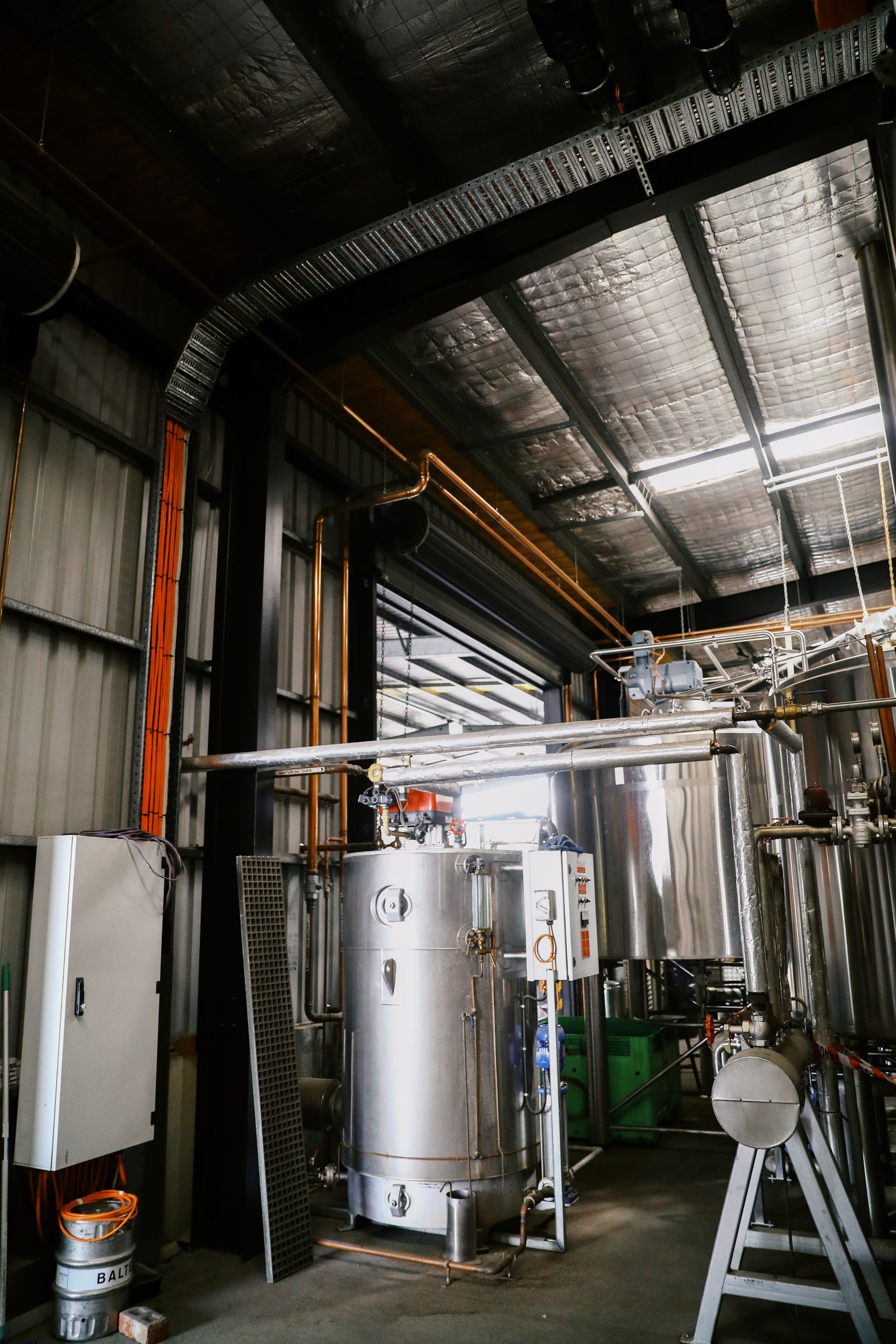 Industrial interior of a brewery featuring stainless steel vats, piping, and reflective insulation on the ceiling.
