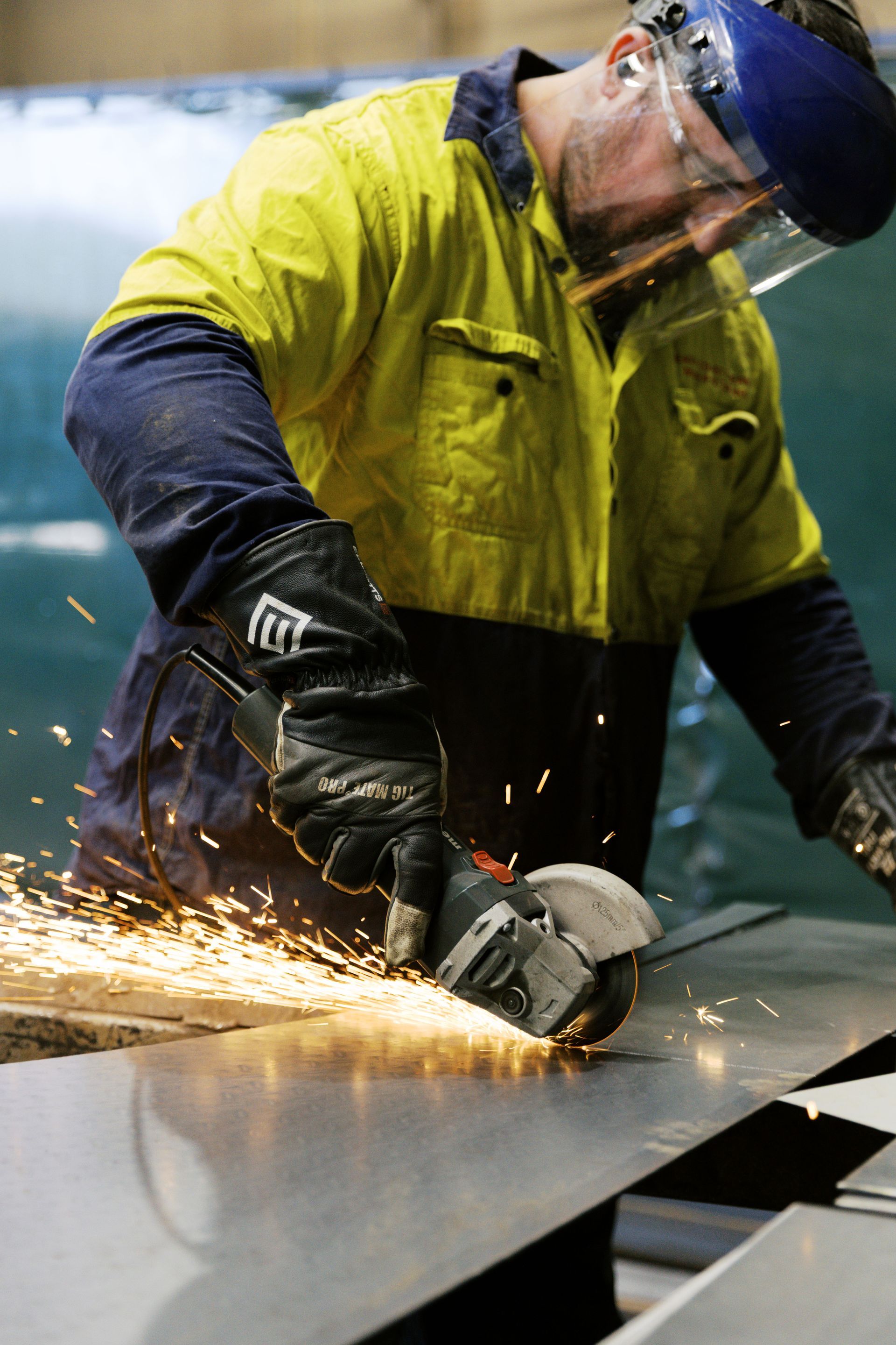 A worker in a high-visibility yellow shirt and face shield uses an angle grinder to cut a metal sheet, creating sparks. A worker in a high-visibility yellow shirt and face shield uses an angle grinder to cut a metal sheet, creating sparks.