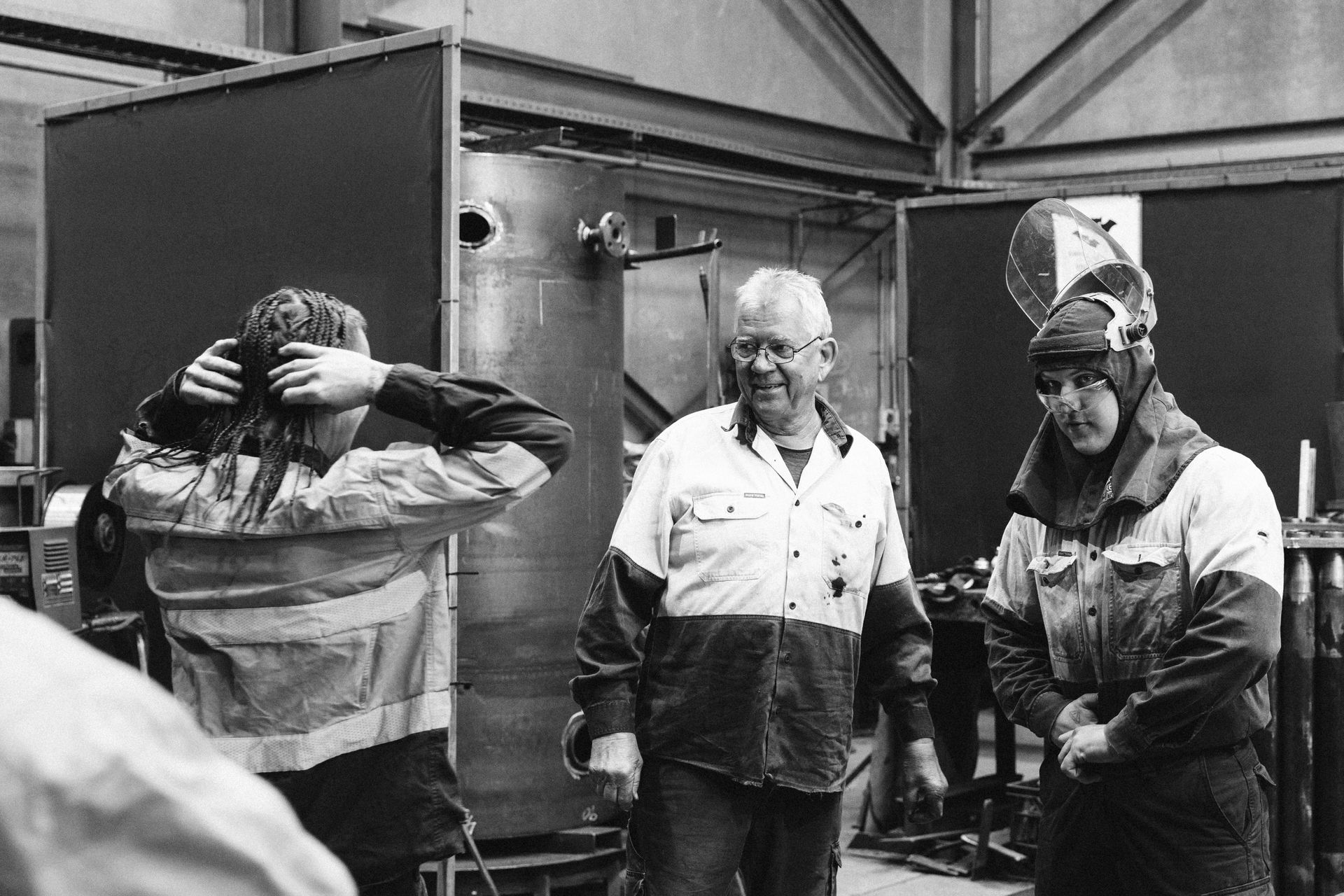 Three people in work uniforms standing in an industrial workshop; one person adjusts their hair, two others converse.