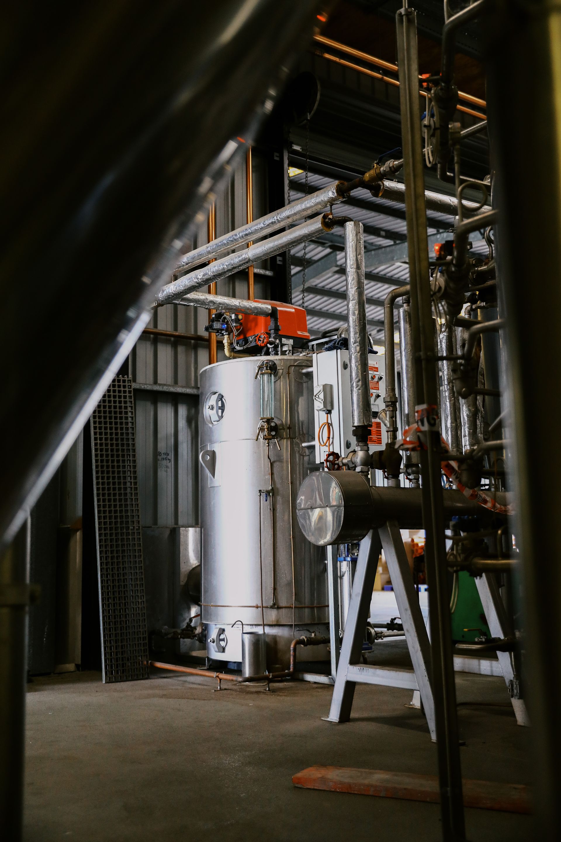 A vertical, silver industrial tank stands on a concrete floor in a workshop, surrounded by pipes and machinery.