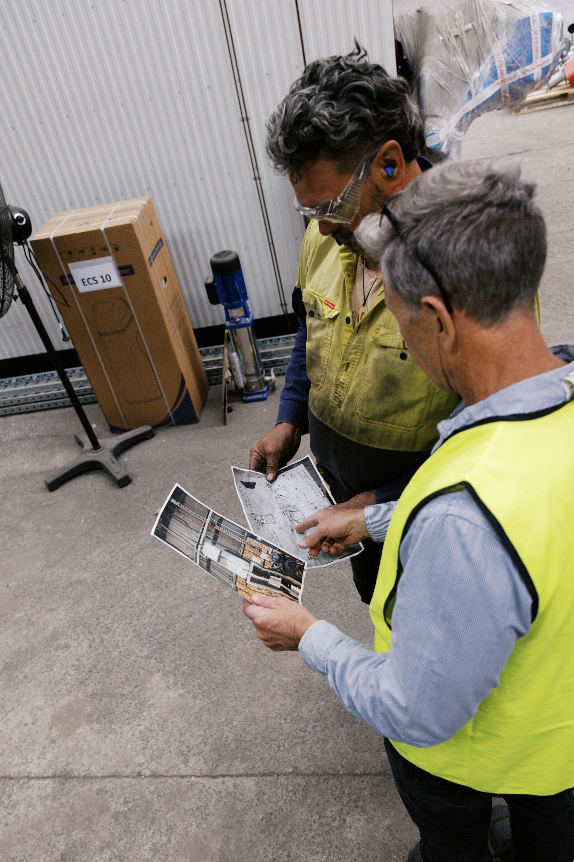 Two workers in high-visibility safety gear study a technical blueprint together in an industrial workspace.