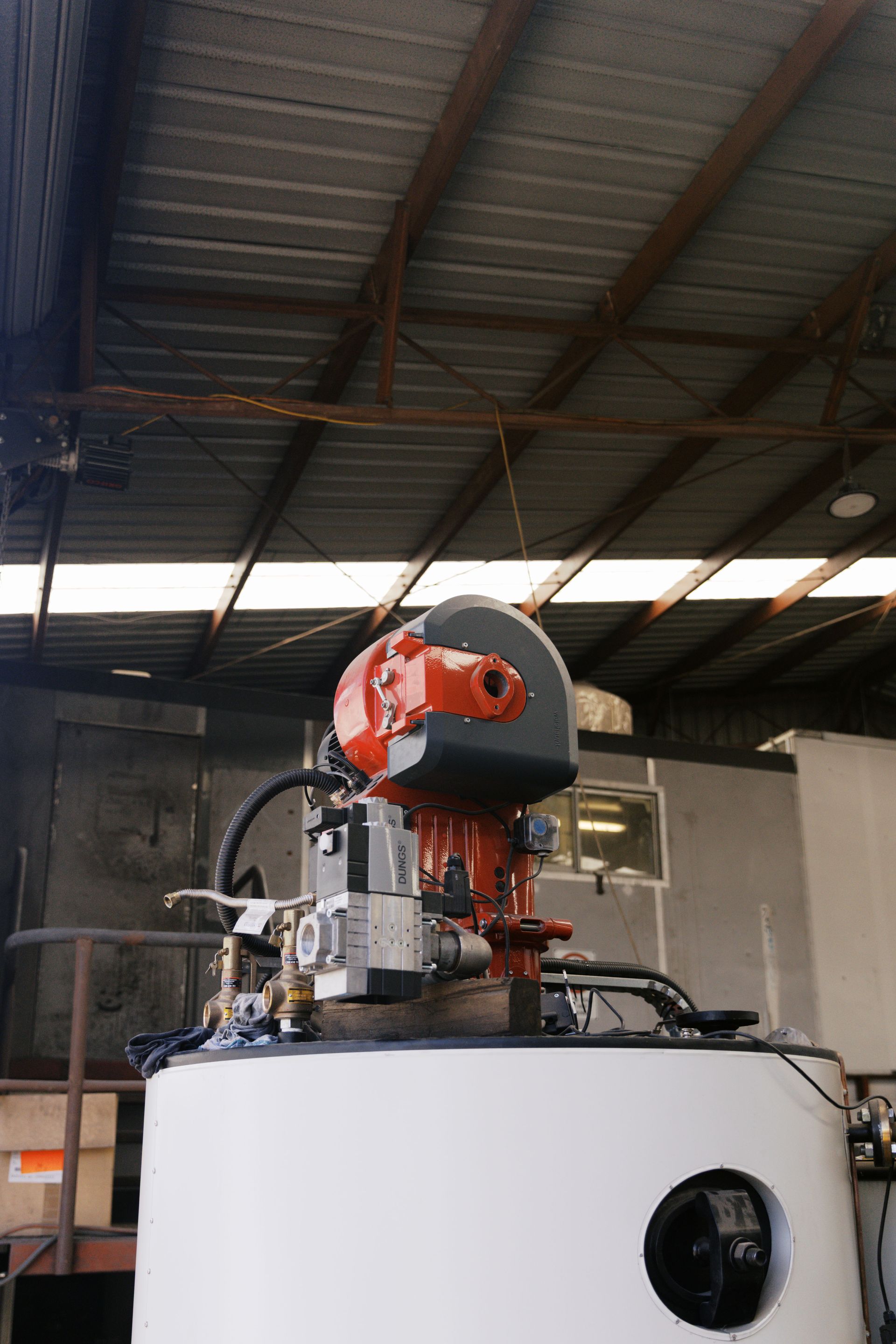 An orange industrial burner mounted on top of a large, white tank inside a warehouse with a corrugated metal ceiling.
