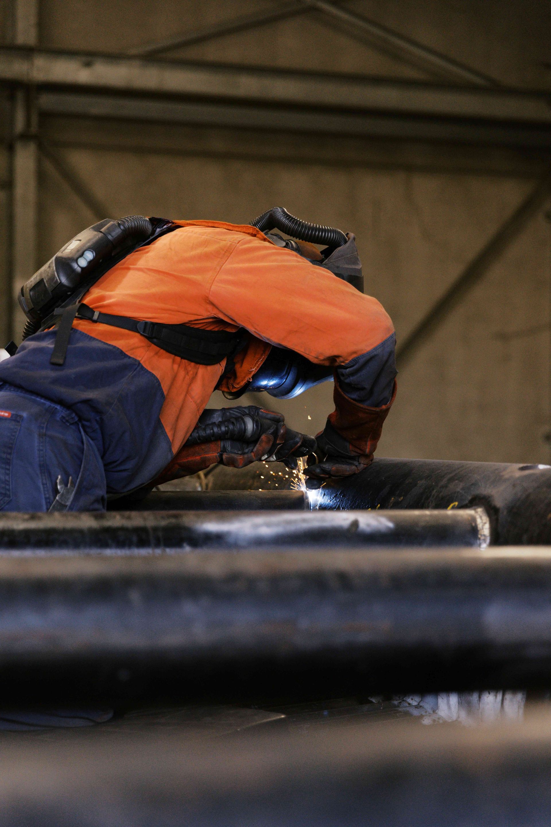 A worker in an orange high-visibility jacket and protective gear welds metal pipes in an industrial workshop. A worker in an orange high-visibility jacket and protective gear welds metal pipes in an industrial workshop.