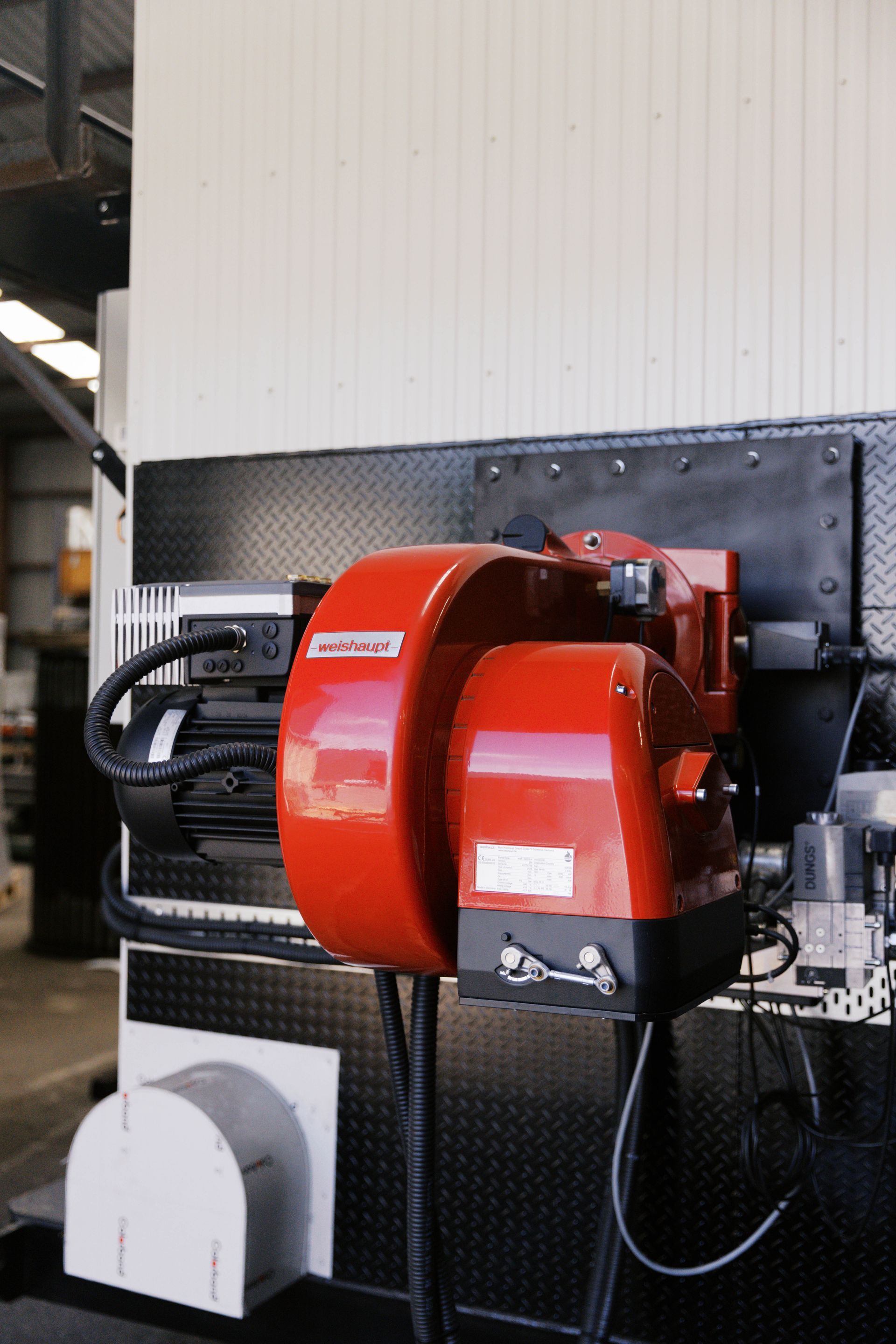 A bright red industrial burner unit mounted on a black diamond-plate wall panel inside a workshop.