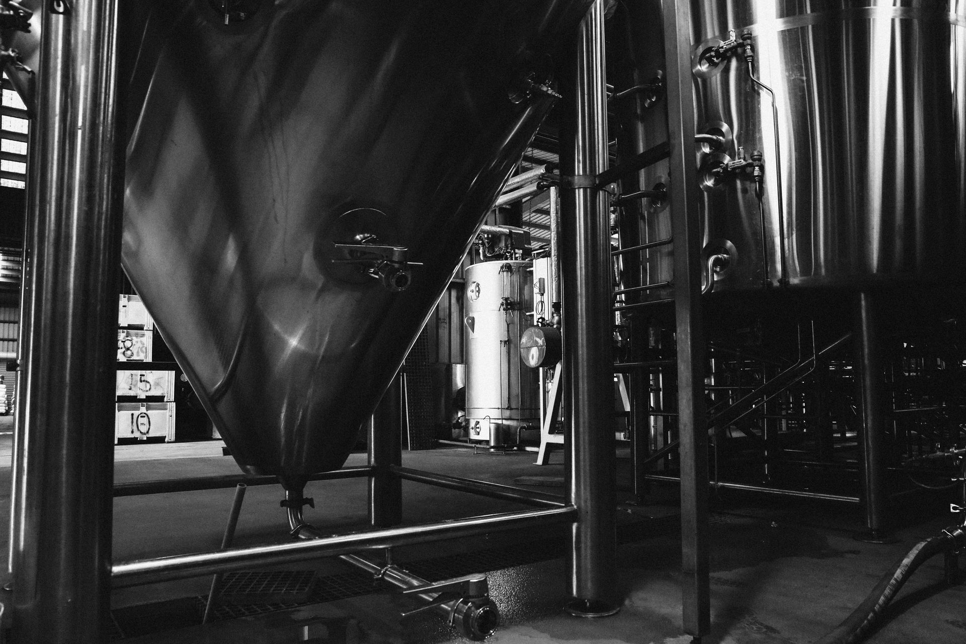 A black and white image of industrial stainless steel fermentation tanks in a brewery.
