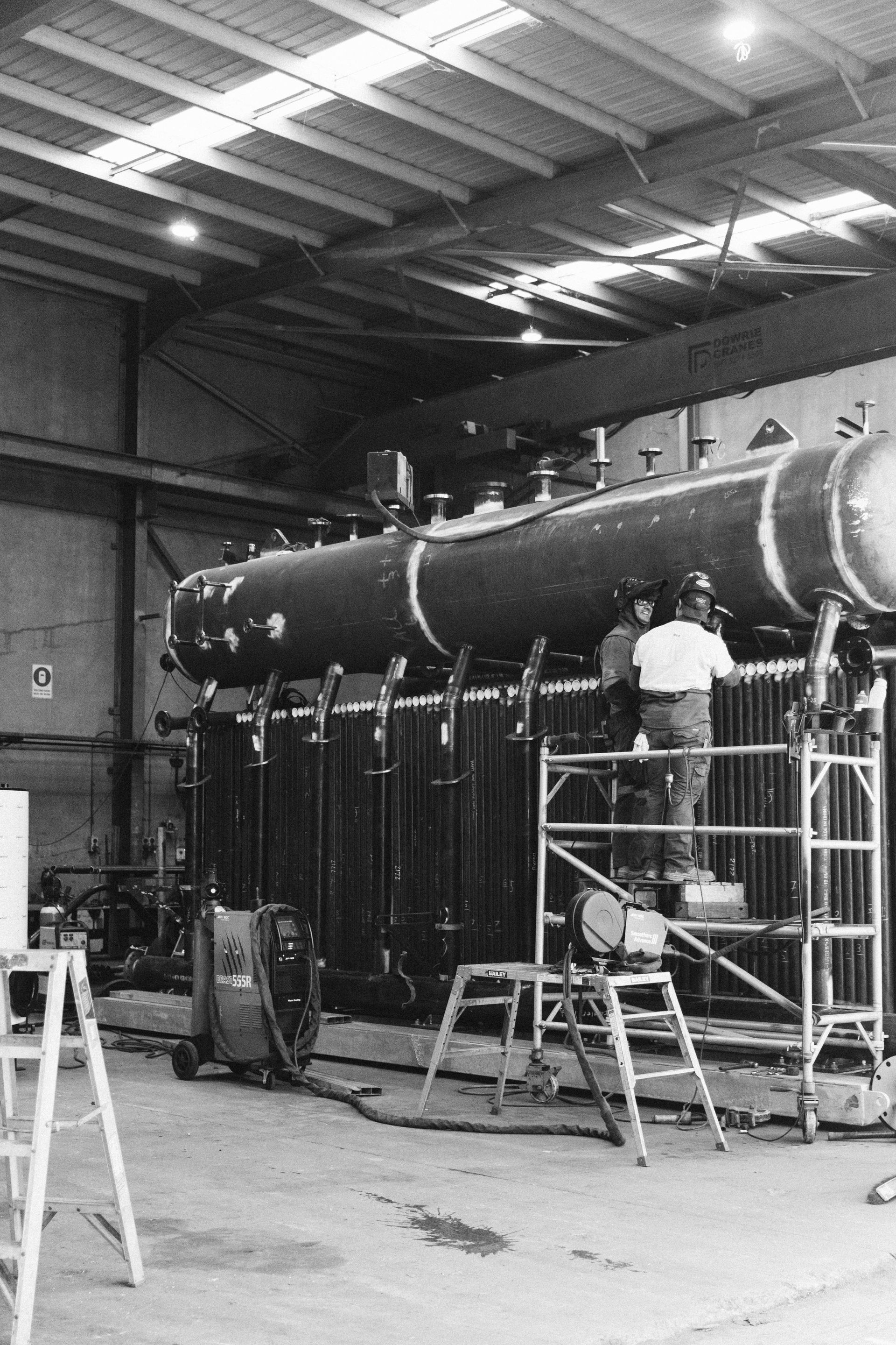 Workers on a scaffold assemble a large industrial boiler in a manufacturing facility.