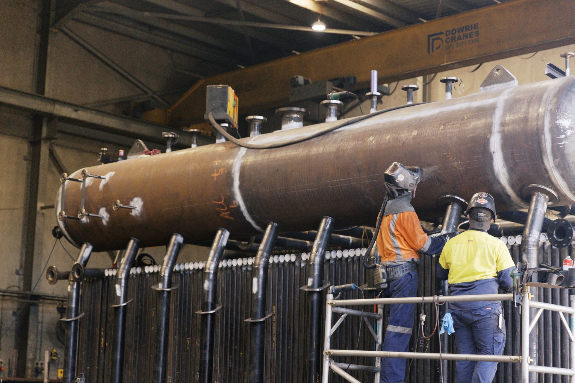 Two workers in safety gear weld a large industrial metal tank in a factory setting. Two workers in safety gear weld a large industrial metal tank in a factory setting.
