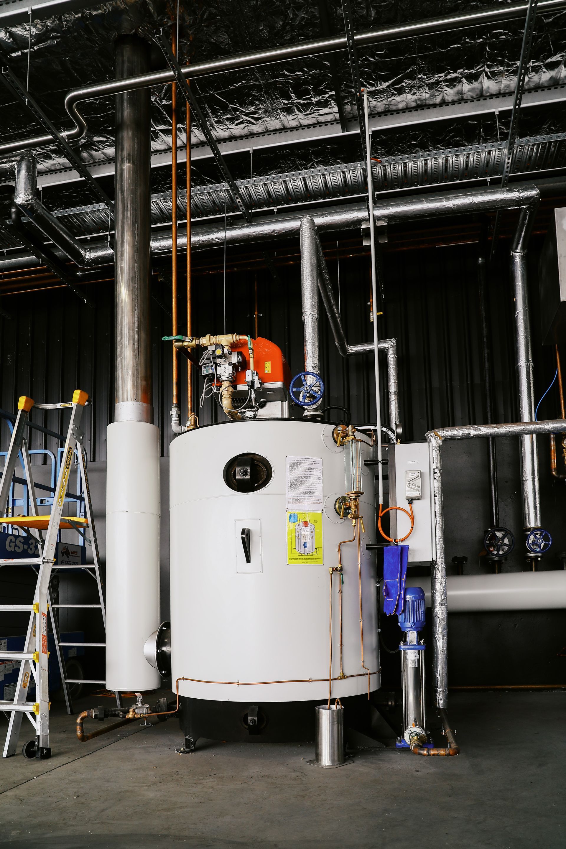 A white vertical industrial boiler stands in a utility room with silver pipes, a tall flue, and a ladder to the left.