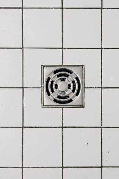 Square metal floor drain centered in white tiled surface.