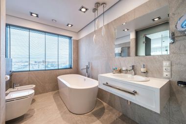 Modern beige bathroom with freestanding tub, vanity sink, toilet, and large window blinds