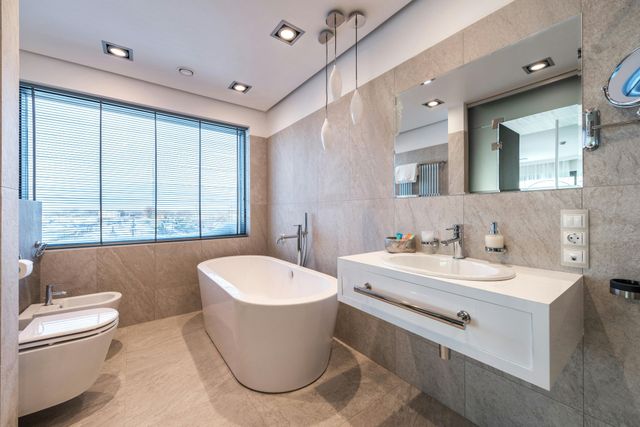 Modern beige bathroom with freestanding tub, vanity sink, toilet, and large window blinds