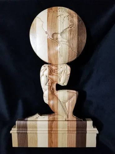 A wooden sculpture of a kneeling figure supporting a globe on their back, set on a multi-toned striped wooden base.