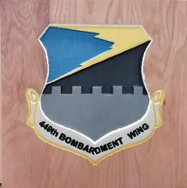 Emblem of the 449th Bombardment Wing: a shield with a yellow and blue lightning bolt over a gray battlement on a wood board.