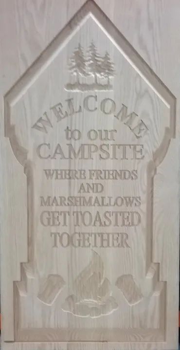 A wooden sign shaped like a tombstone, carved with pine trees, a campfire, and the text: