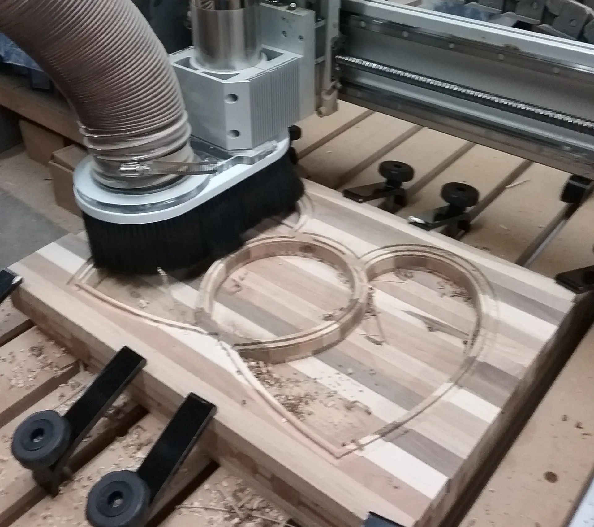 A CNC machine with a dust collection boot cuts a heart shape into a striped wooden board held by clamps on a workbench.