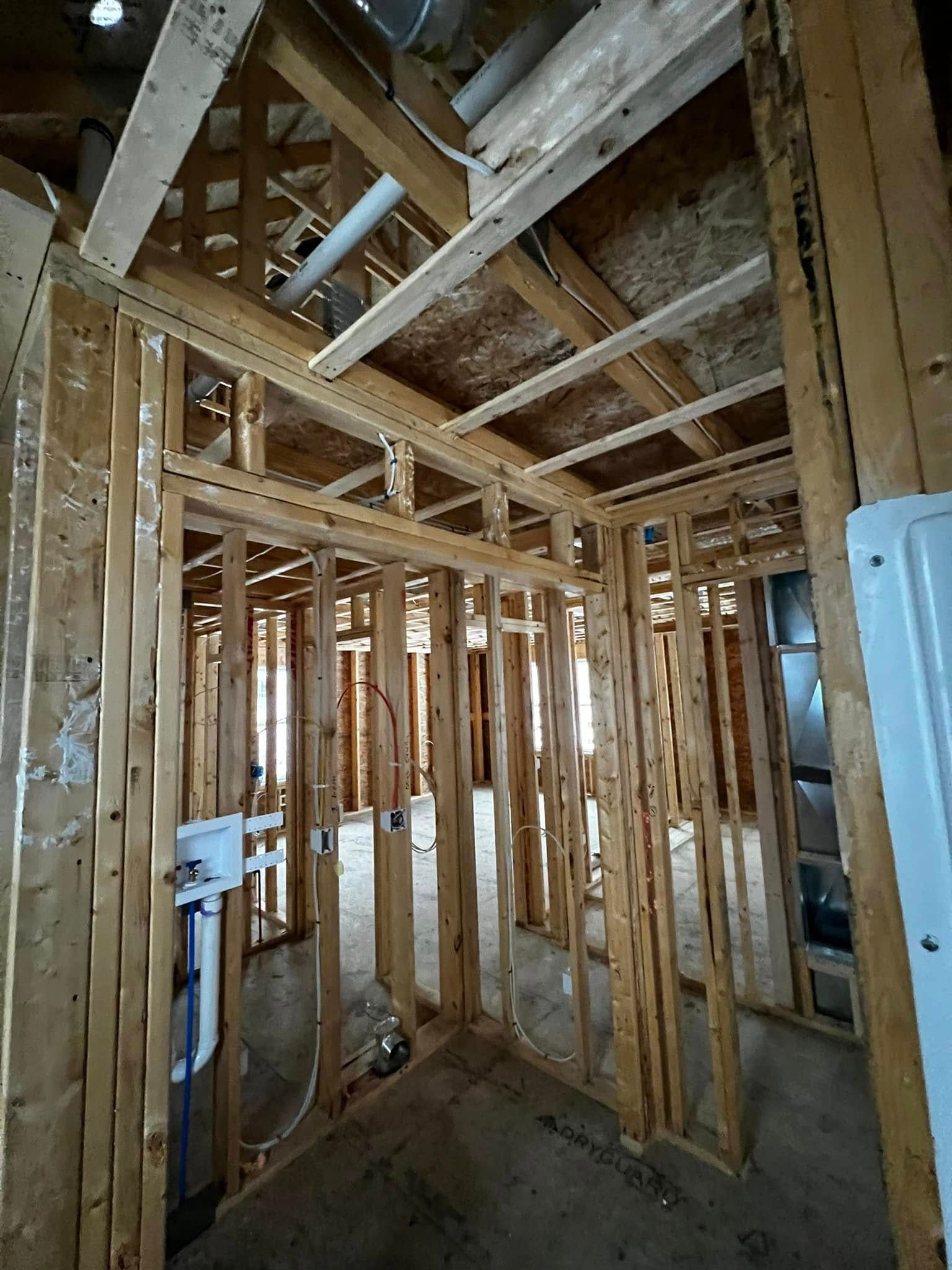 Interior view of a building's wooden frame under construction. Visible studs, beams, pipes, and wiring.