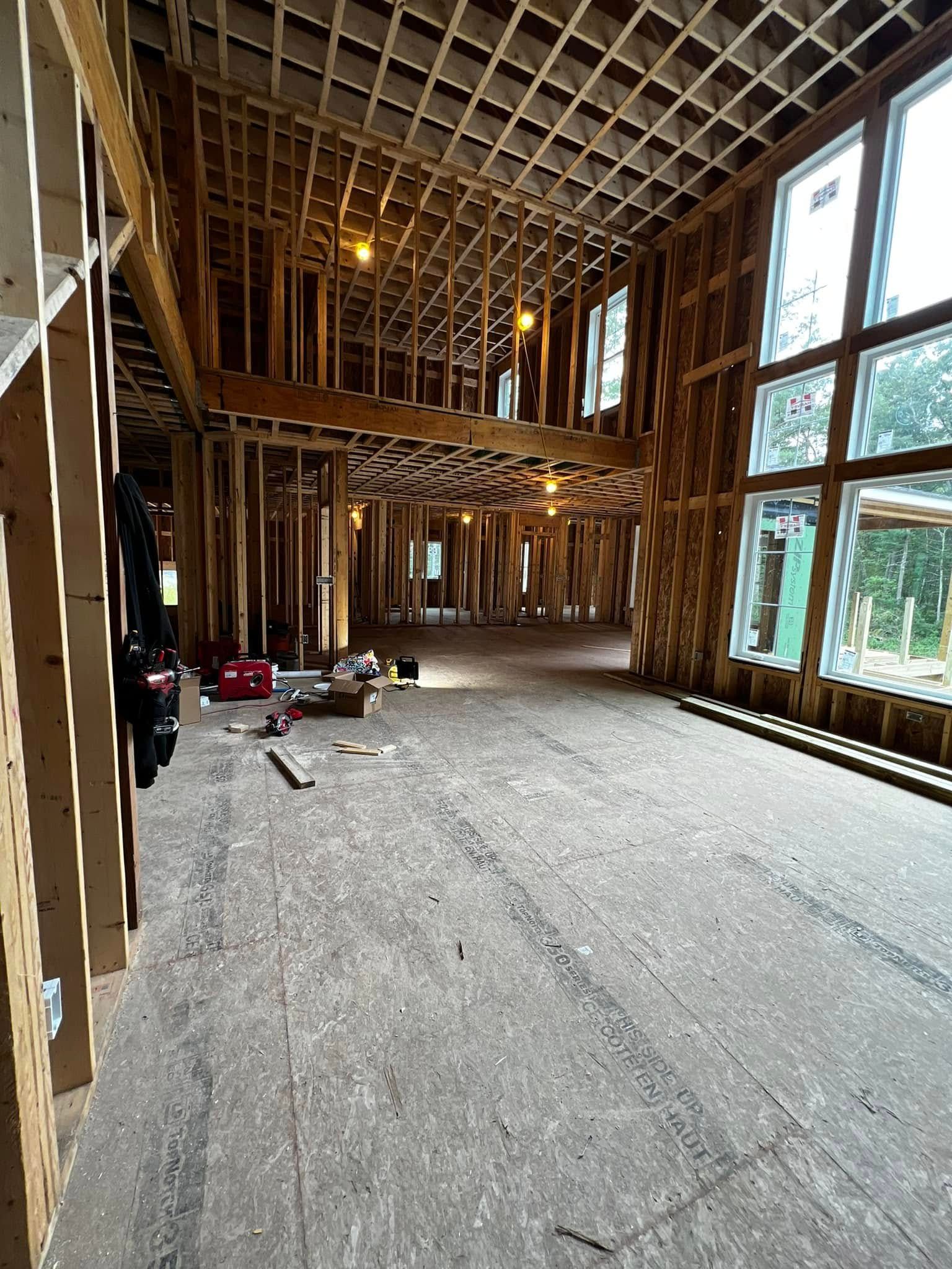 Interior view of a building under construction, showing wooden framing, unfinished floors, and large windows.