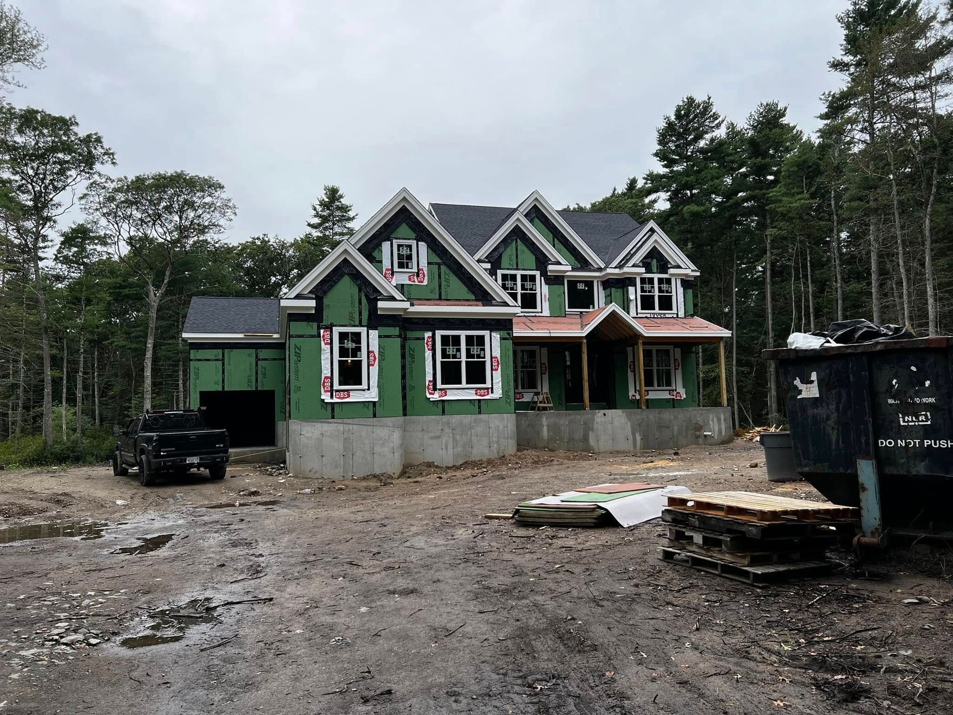 A two-story house under construction surrounded by trees; green siding, gray roof, and cloudy sky.