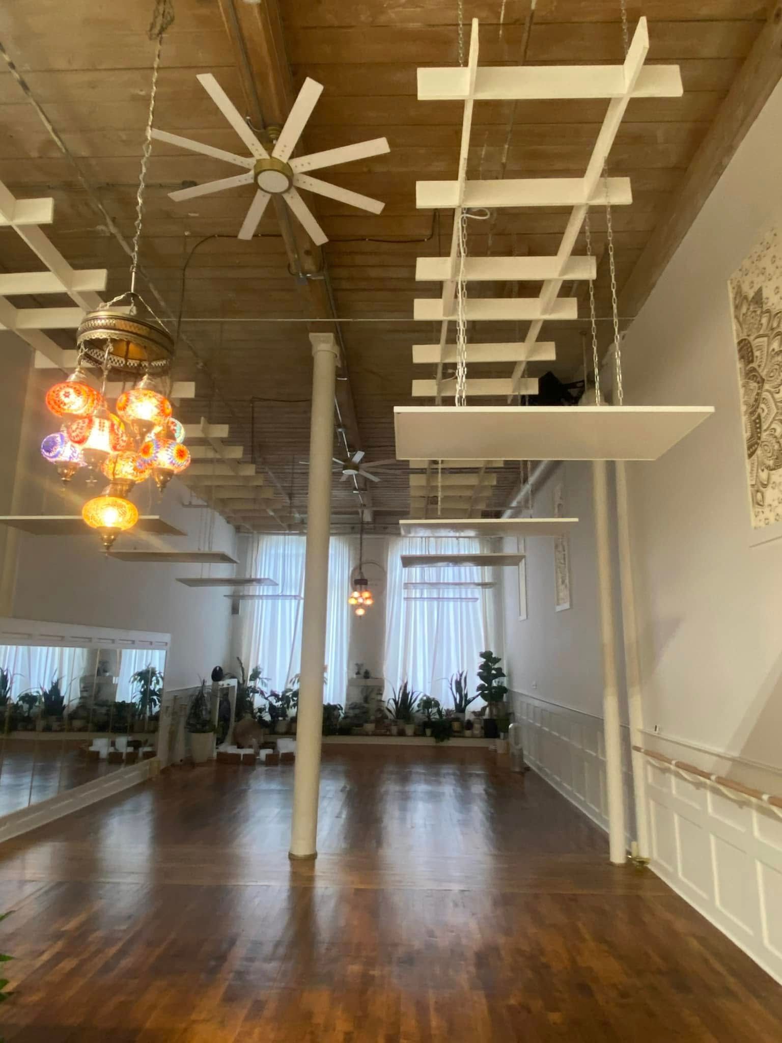 Spacious studio with wood floors, plants, mirrors, white pillars, and hanging light fixtures.