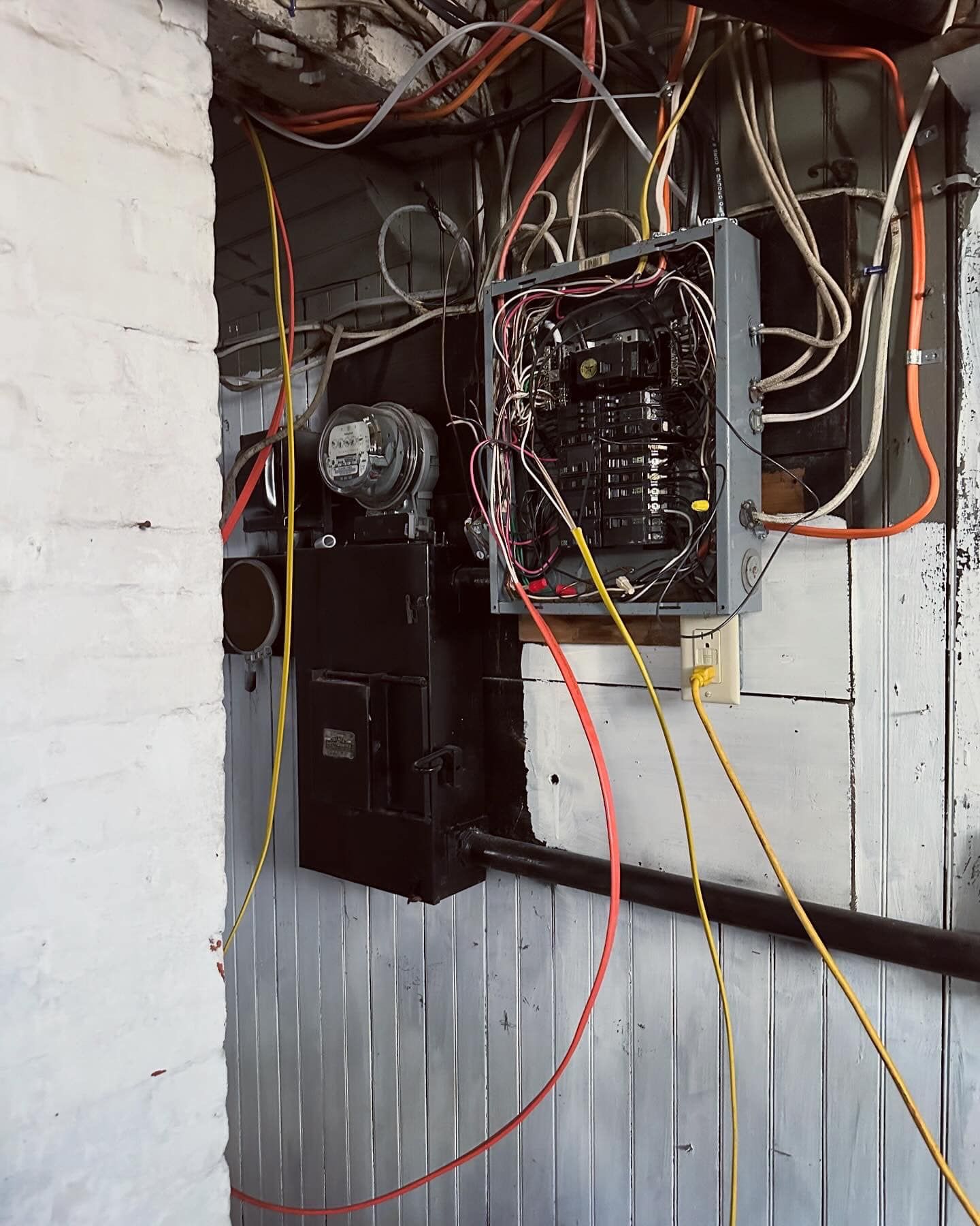 Electrical panel with exposed wires in a basement, orange and yellow wires against a gray wall.