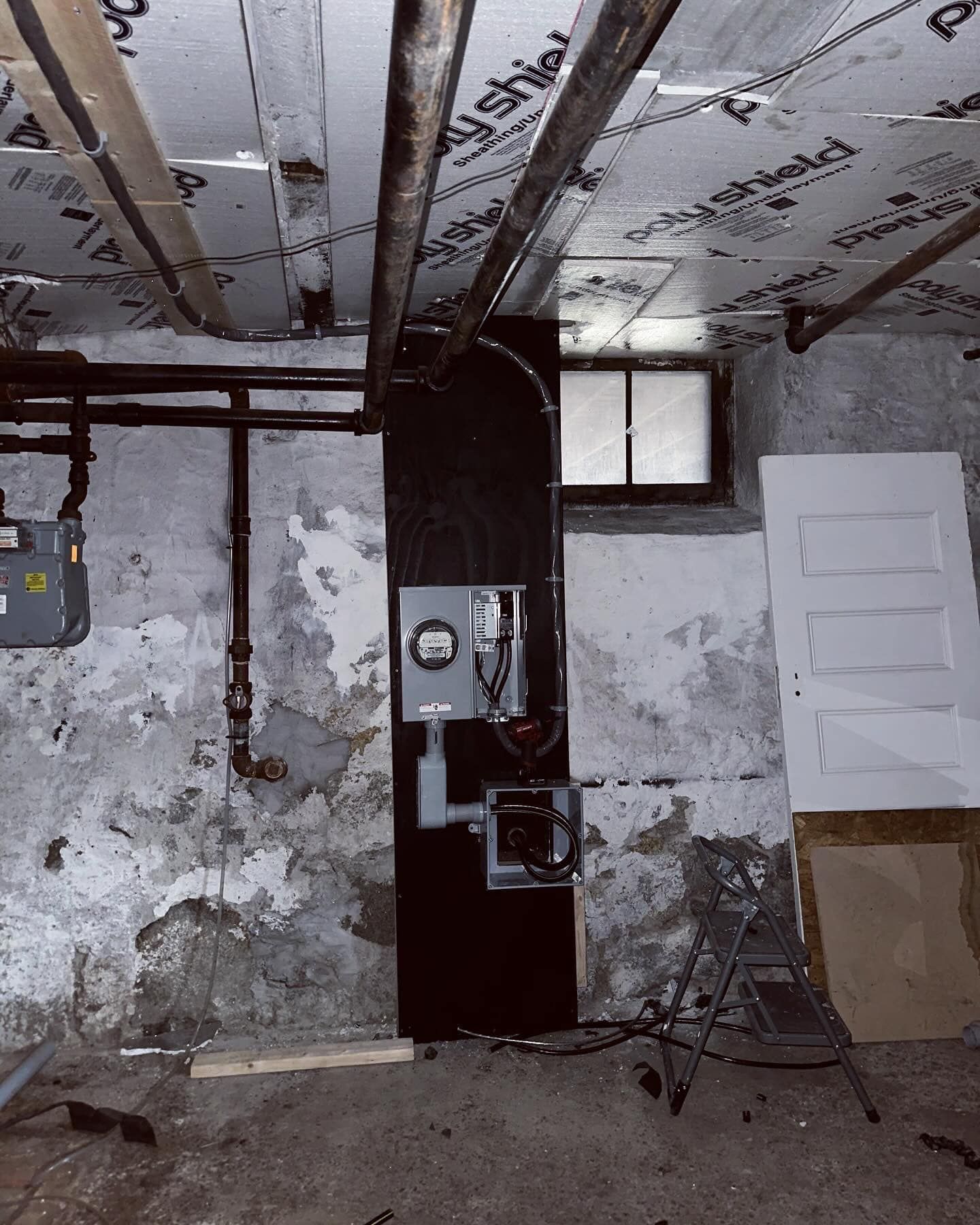 Basement with electrical panel, pipes, and window. White door and gray meter on wall.