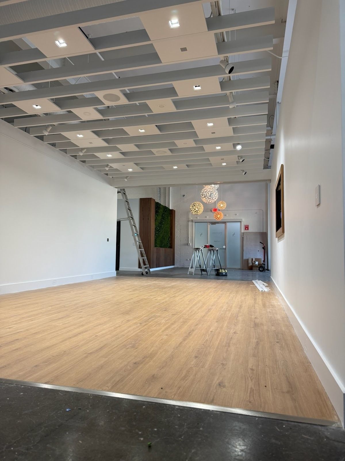 Empty gallery space with light wood floor, white walls, and open ceiling. A ladder and artwork are visible.