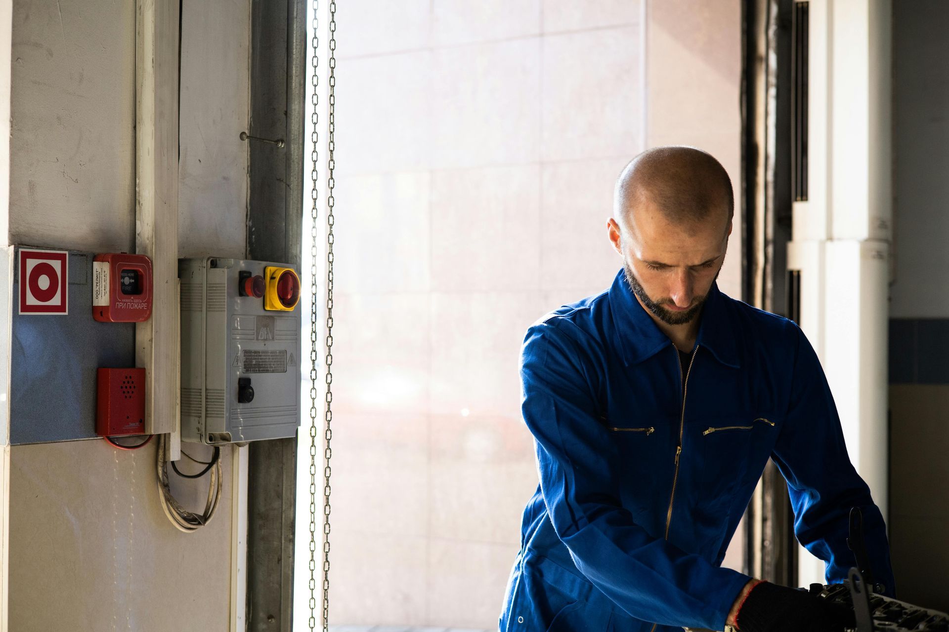 Mechanic in blue jumpsuit works near open door with machinery controls.