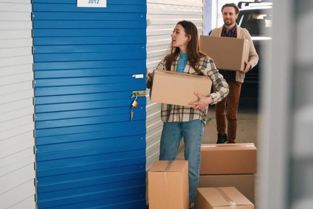 A couple carrying large cardboard boxes and getting them into a blue-door storage unit.