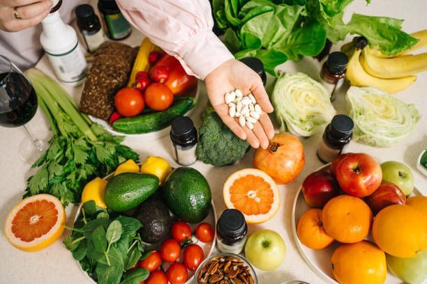 A person is holding pills in their hand in front of a table filled with fruits and vegetables.