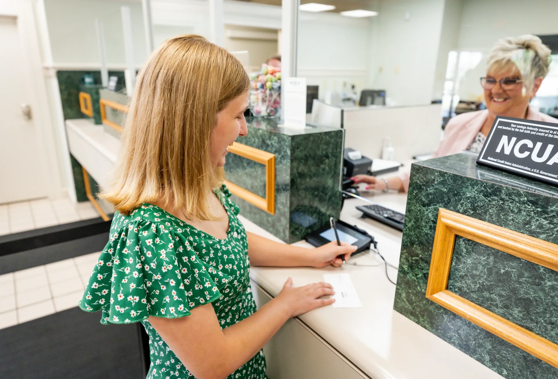 Young girl signing a document at a bank counter, smiling at a teller.