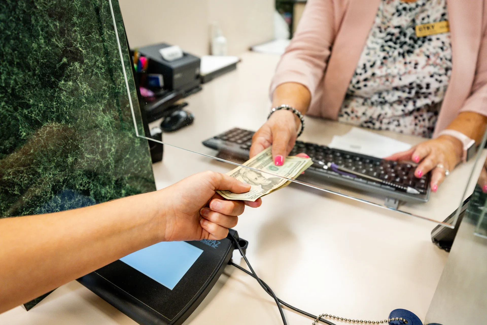 Hand exchanging cash with a bank teller at a counter with a computer and protective screen.