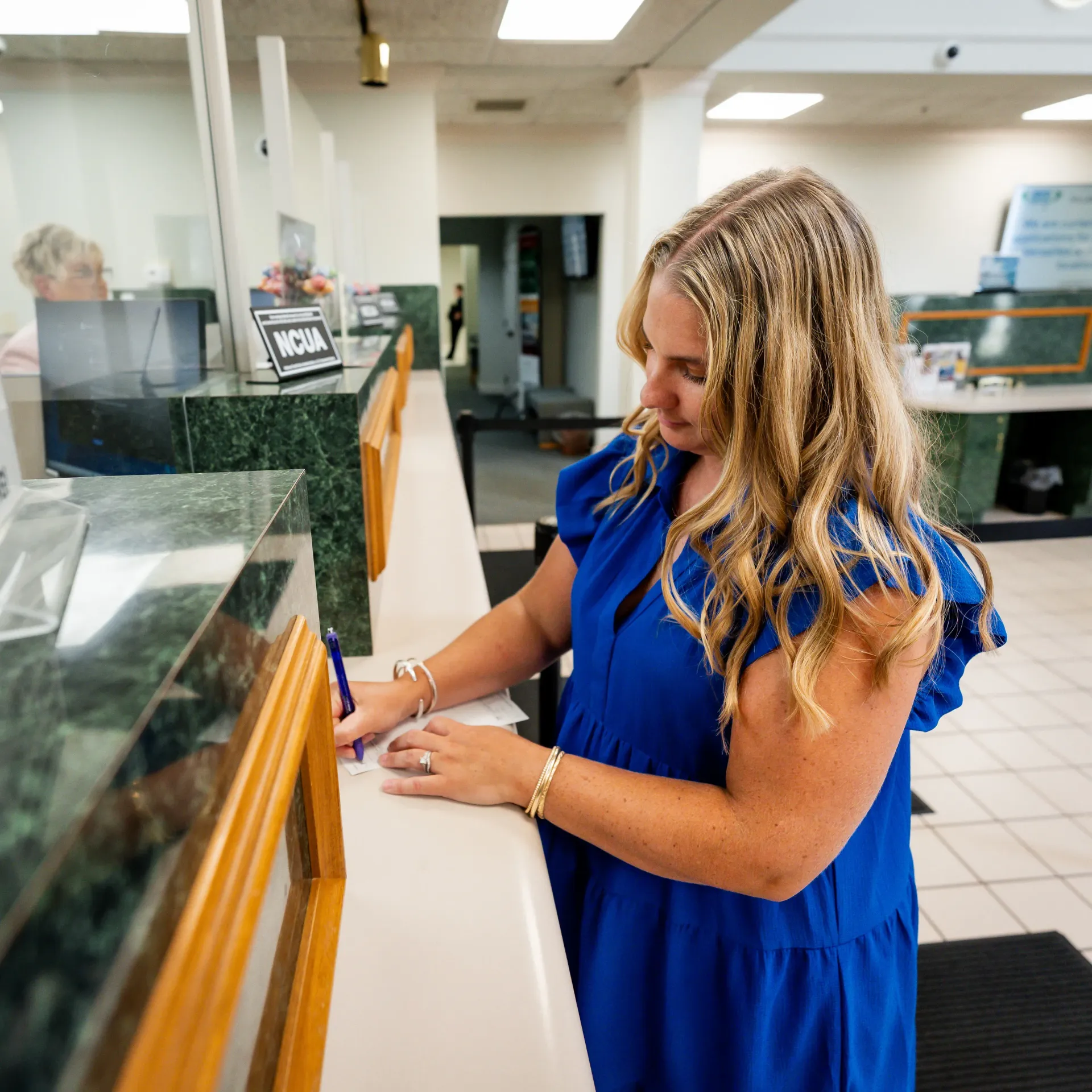 Woman in blue dress writing at bank counter.