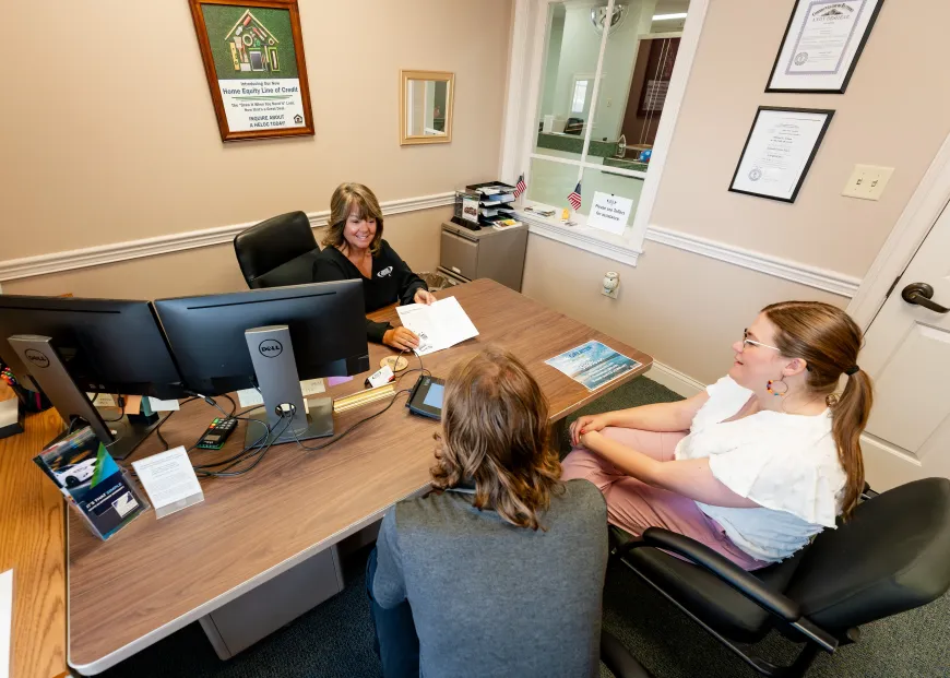 Three people at desk in office; woman at computer, two clients.