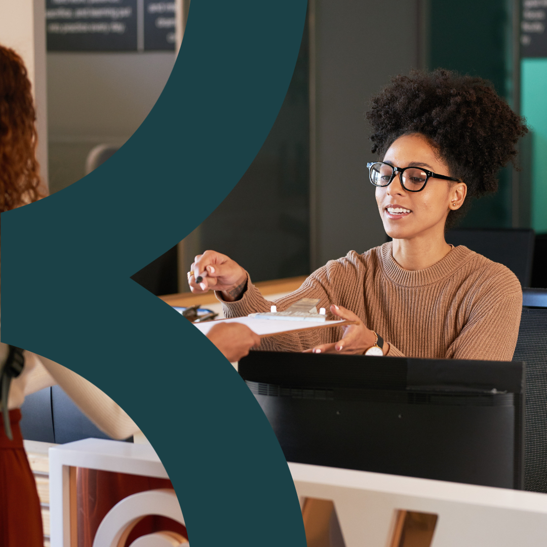 Woman at a reception desk assisting a visitor with paperwork.