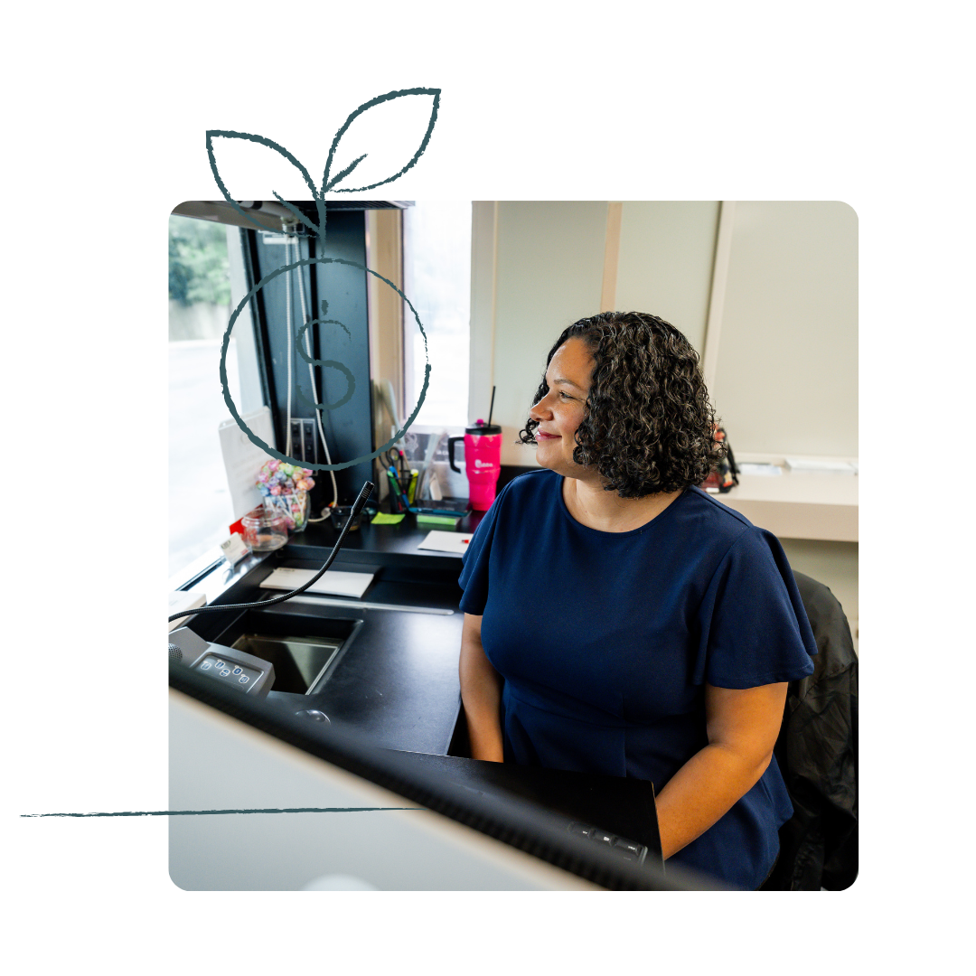 Woman at desk, looking to the side. Blue dress, office setting, dark hair.