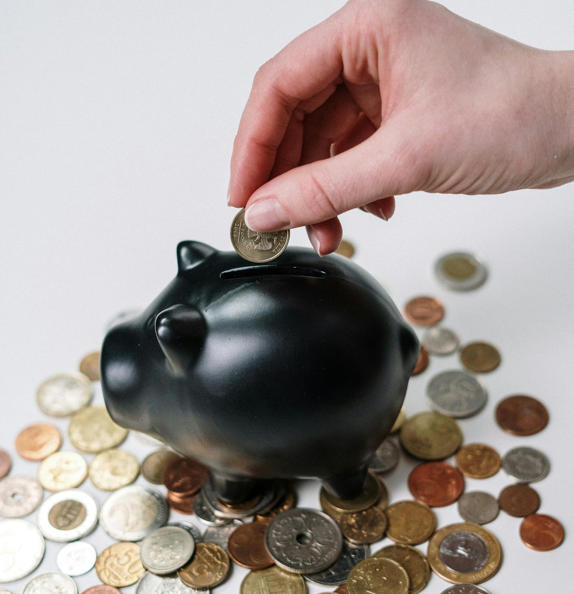 Hand inserting coin into a black piggy bank, surrounded by loose change.