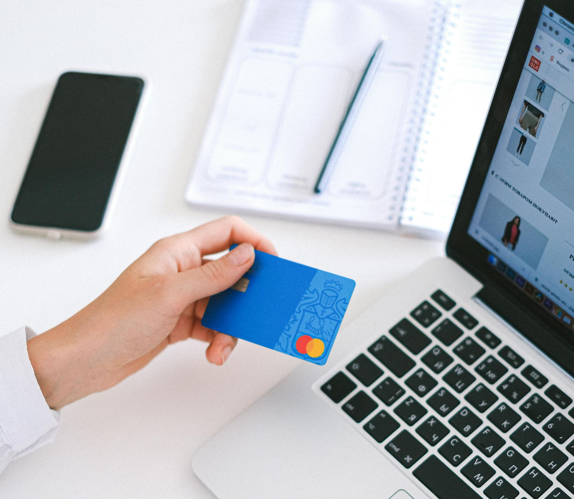 Hand holding a blue credit card near a laptop, notebook, and smartphone on a white surface.