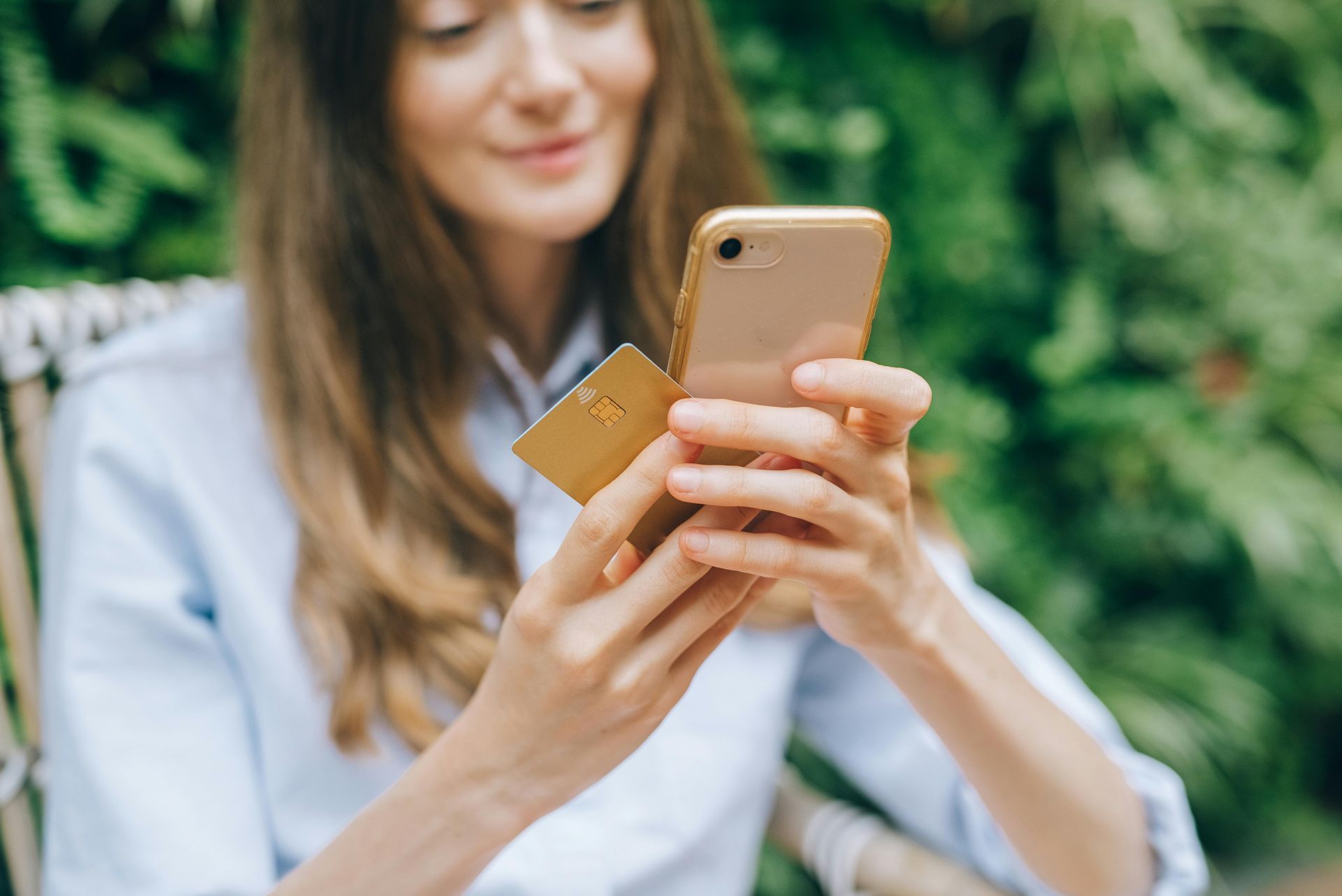 Woman holding gold credit card and smartphone, smiling outdoors near greenery.