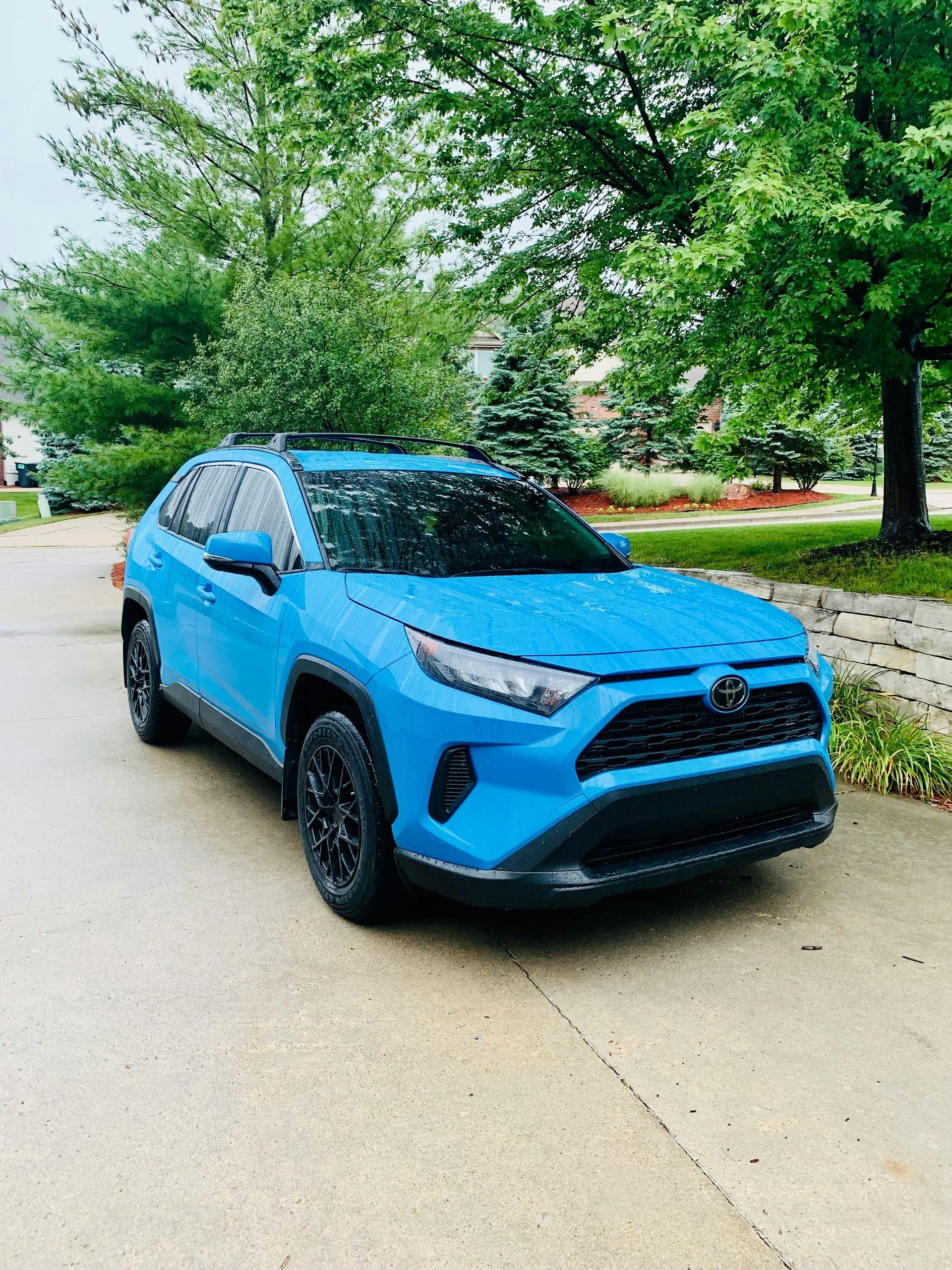 Blue Toyota RAV4 SUV parked on a paved road with black wheels, trees in the background.
