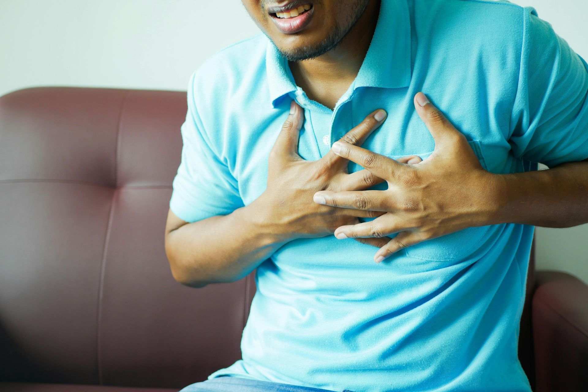 A man in a blue shirt is sitting on a couch holding his chest.